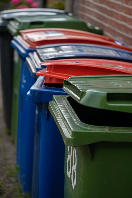 A row of large, plastic wheelie bins lined up on a pavement alongside a brick wall, with the front bin being green and the others behind it in blue, red, and green colours. The bins are used for waste