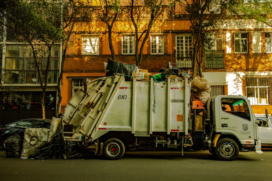 A row of large, plastic wheelie bins lined up on a pavement alongside a brick wall, with the front bin being green and the others behind it in blue, red, and green colours. The bins are used for waste