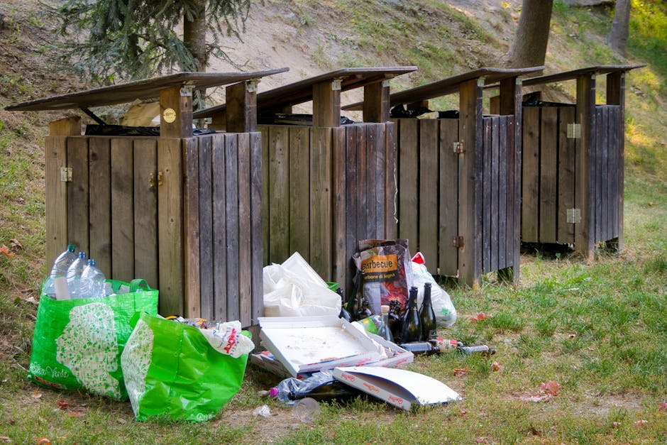 Four wooden outdoor rubbish and recycling bins with sloped lids are arranged in a row on a grassy area, surrounded by trees and sloping terrain in the background. The bins are made of weathered, dark-